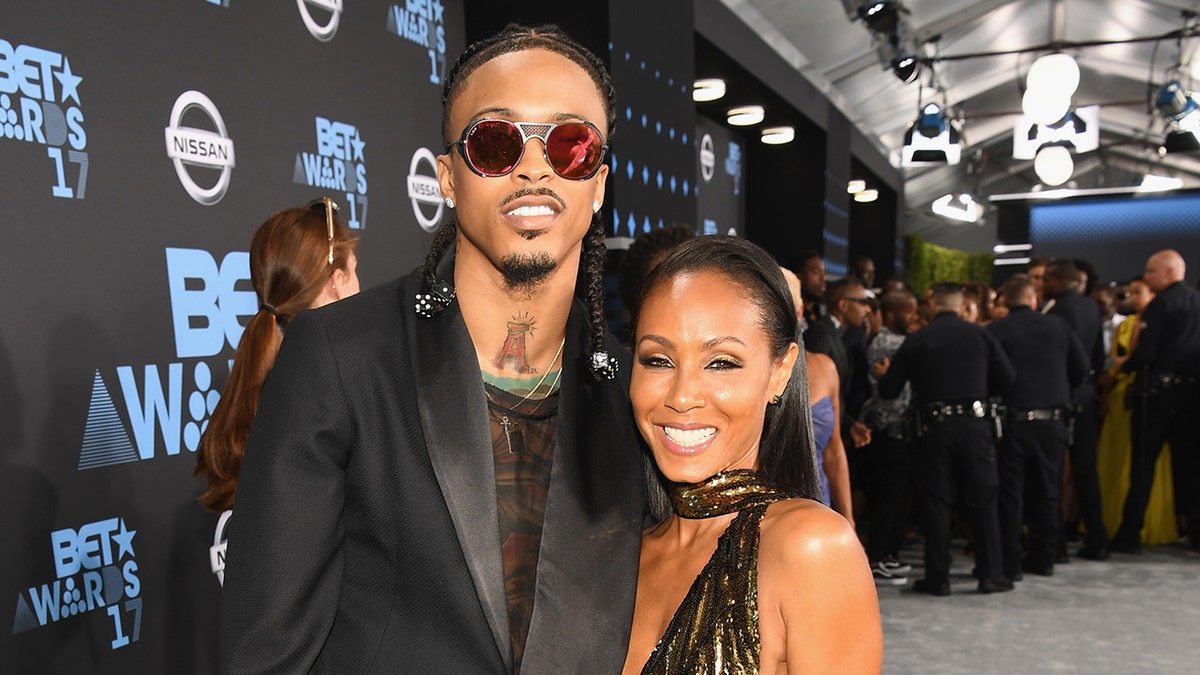 August Alsina y Jada Pinkett Smith sonriendo mientras ambas visten atuendos negros