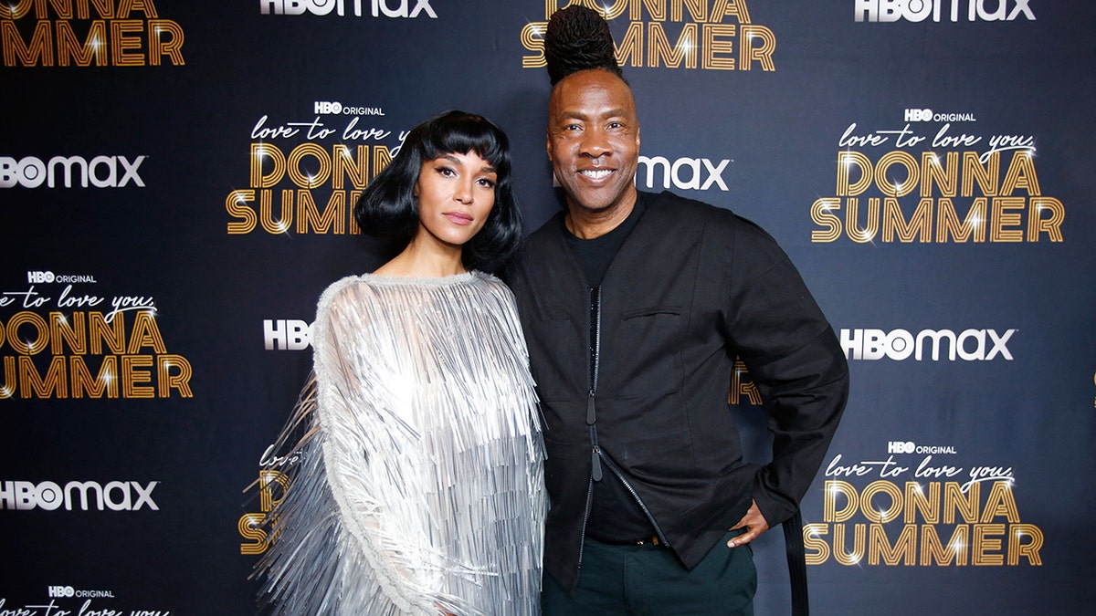 Brooklyn Sudano con un vestido plateado posando junto a Roger Ross Williams con un conjunto completamente negro