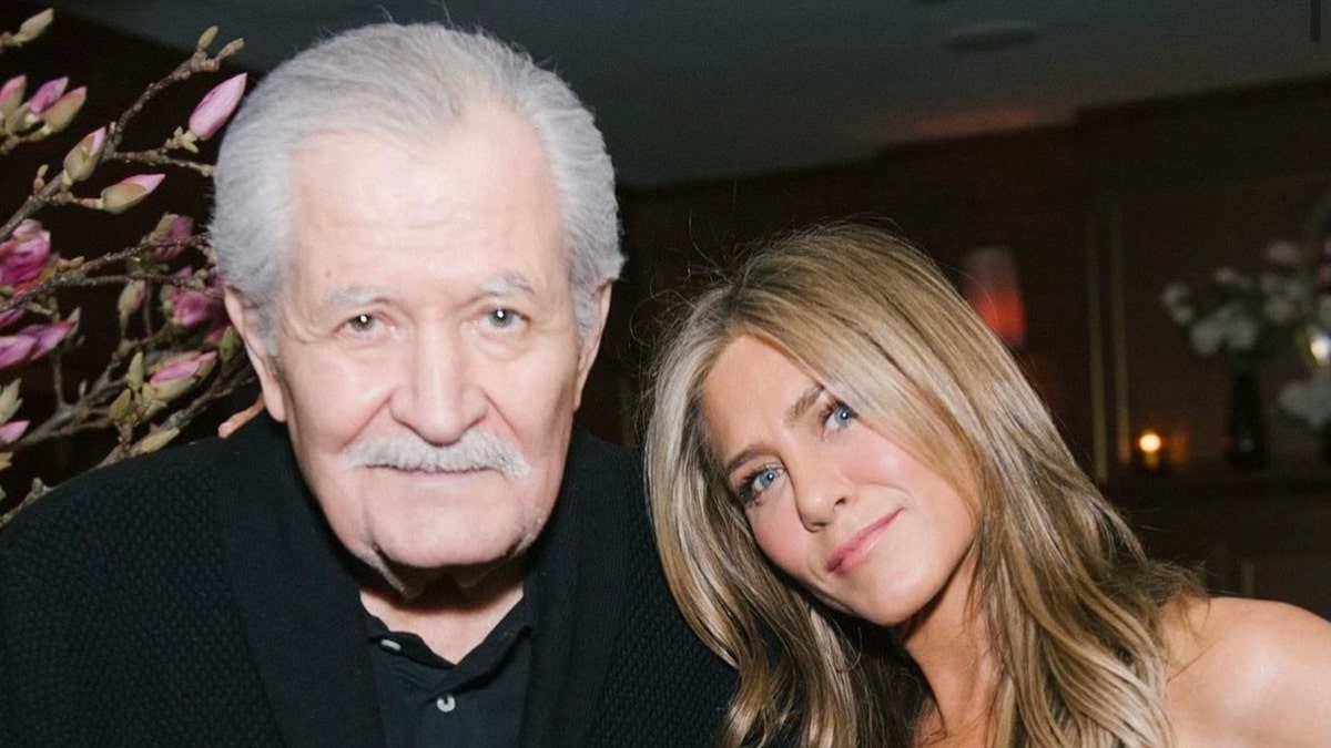 Jennifer Aniston con un atuendo negro inclina la cabeza hacia father John, también en negro