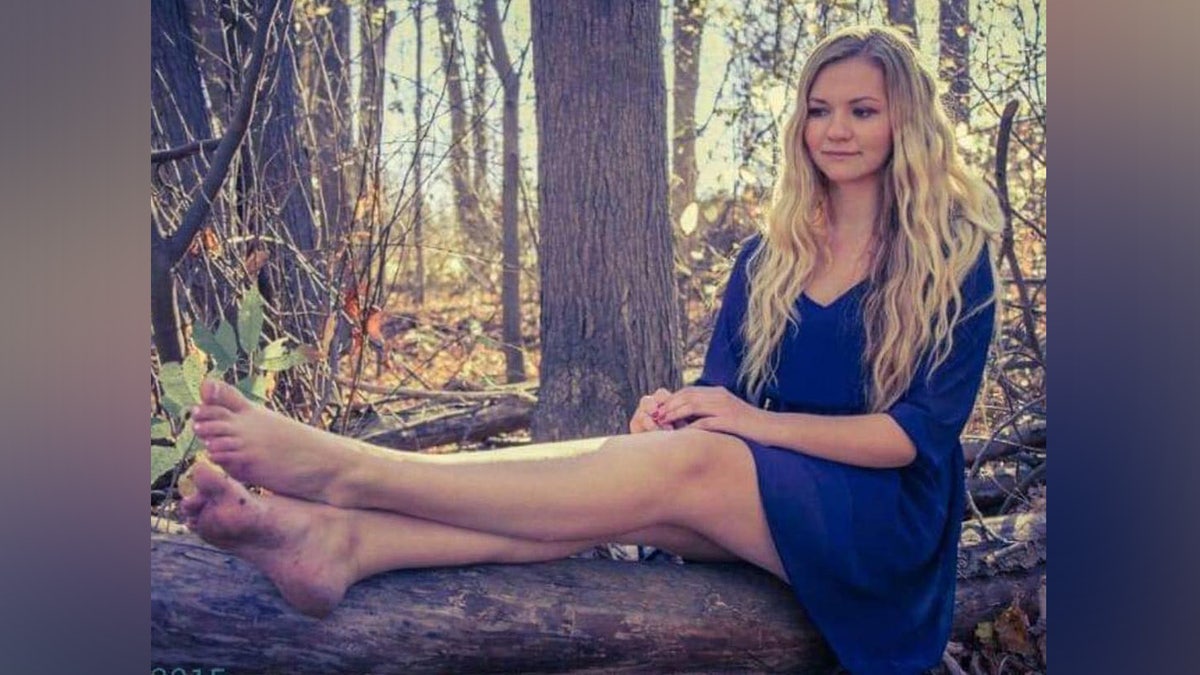 Gina con un vestido azul y sentada en un tronco en el bosque.