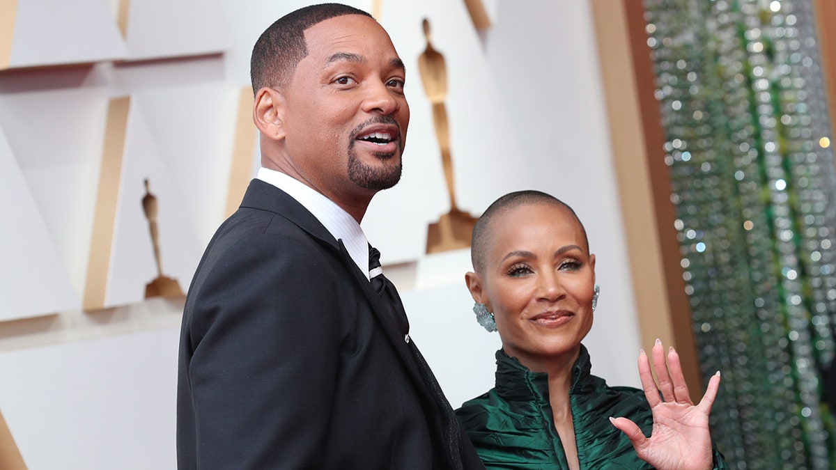 Will Smith mira ligeramente hacia arriba en la alfombra de los Oscar con Jada Pinkett Smith sonriendo a su lado con un vestido verde